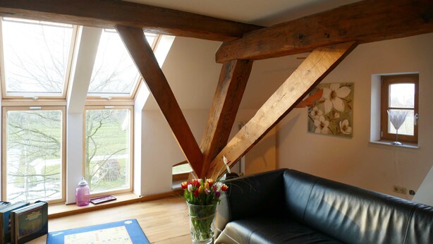 Ein Zimmer mit einer schwarzen Ledersofa, Holzbalken, großen Fenstern und einer Blumenvase.