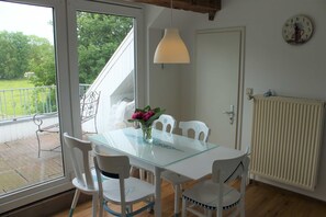 Un comedor con una mesa de vidrio, sillas blancas, un florero con flores, una lámpara colgante y vistas a un balcón con barandilla.