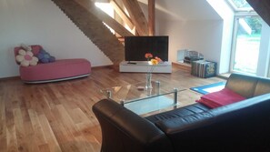 Una sala moderna con piso de madera, un sofá rosa, un sofá de cuero negro, una mesa de centro de vidrio, un televisor de pantalla plana y una escalera con vigas de madera.