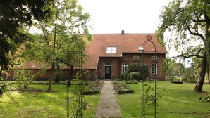 Ein Backsteinhaus mit rotem Ziegeldach, einem Betonweg, der zum Eingang führt, und einem gepflegten Garten mit Bäumen und Sträuchern.