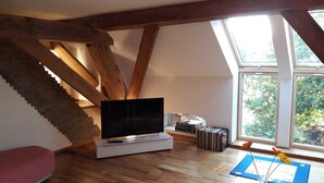 Ein Zimmer mit Fernseher auf einem Schrank, Holzbalken, einer Treppe und einem Fenster mit Blick auf Bäume.