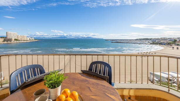 Un balcon avec une table en bois, deux chaises, une plante en pot et un saladier rempli d’oranges. La vue comprend une plage et l’océan.