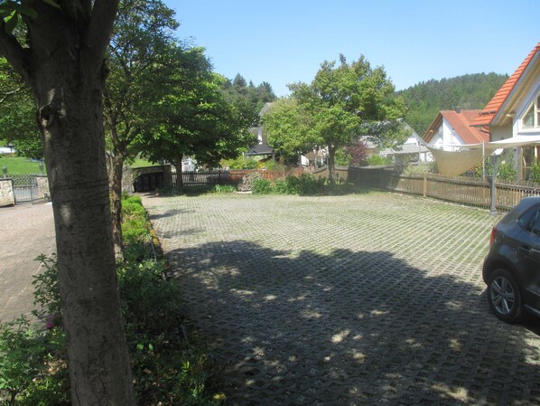 A paved courtyard with a car parked on the right, surrounded by trees and a fence.