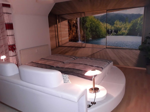 A modern bedroom with a large bed, a bedside table, a lamp, and a large window with a scenic view.
