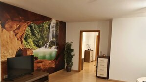 Una sala de estar con un televisor sobre un mueble de madera, un sofá, un tapete estampado, una planta en maceta y una pared con un mural de cascada.