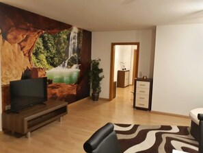 Una sala de estar con un televisor sobre un mueble de madera, un sofá, un tapete estampado, una planta en maceta y una pared con un mural de cascada.