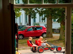 On aperçoit, à travers une fenêtre, deux enfants à bord d’un petit véhicule, avec une voiture rouge garée à l’extérieur.