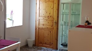 A room with a wooden door, a glass cabinet with dishes, a small trash bin, and a window with a plant.