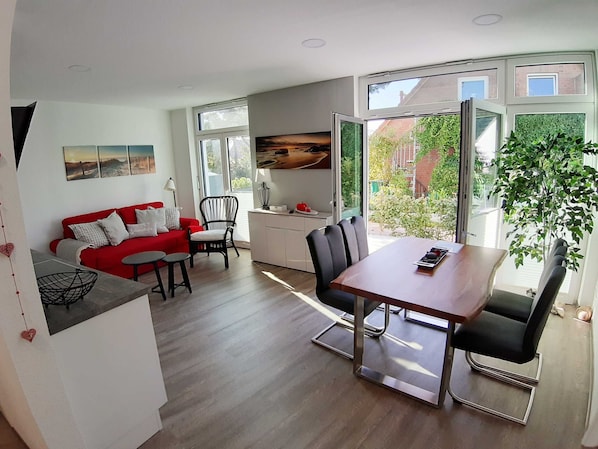 Un salon moderne avec un canapé rouge, une table à manger et des chaises, un coin cuisine et de grandes fenêtres donnant sur l’extérieur.