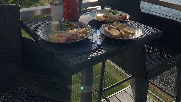 Een eettafel met borden eten, een rode mok en een glas water op een balkon met uitzicht op de oceaan.