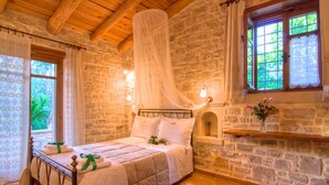 Une chambre avec un mur en pierre, un plafond en bois, un lit avec du linge de lit blanc, une table de chevet, une fenêtre avec des rideaux et une étagère murale décorée de fleurs.