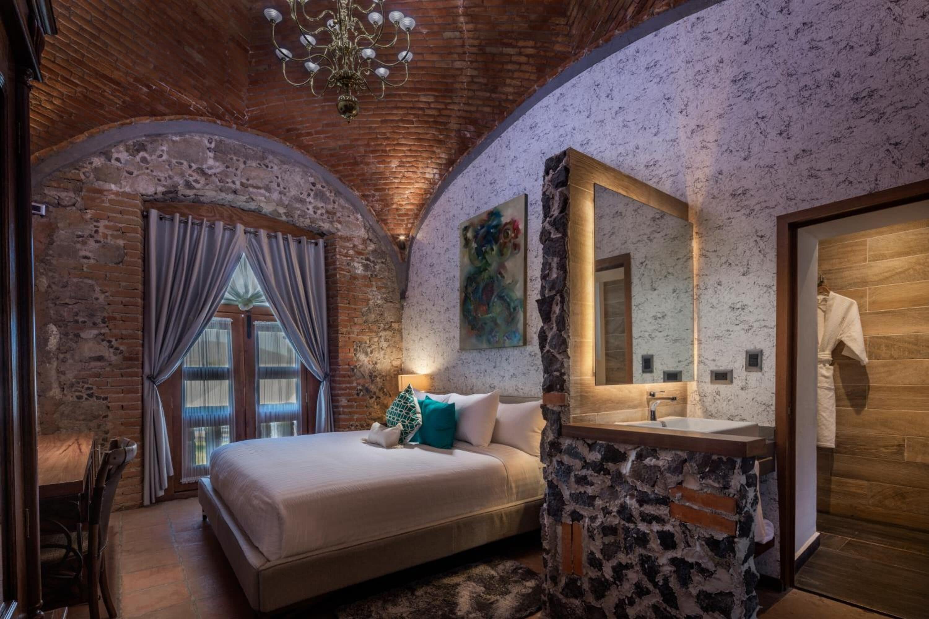 Camera da letto con un letto grande, una parete decorata in pietra, una porta in legno e un bagno con uno specchio ampio e un lavandino.