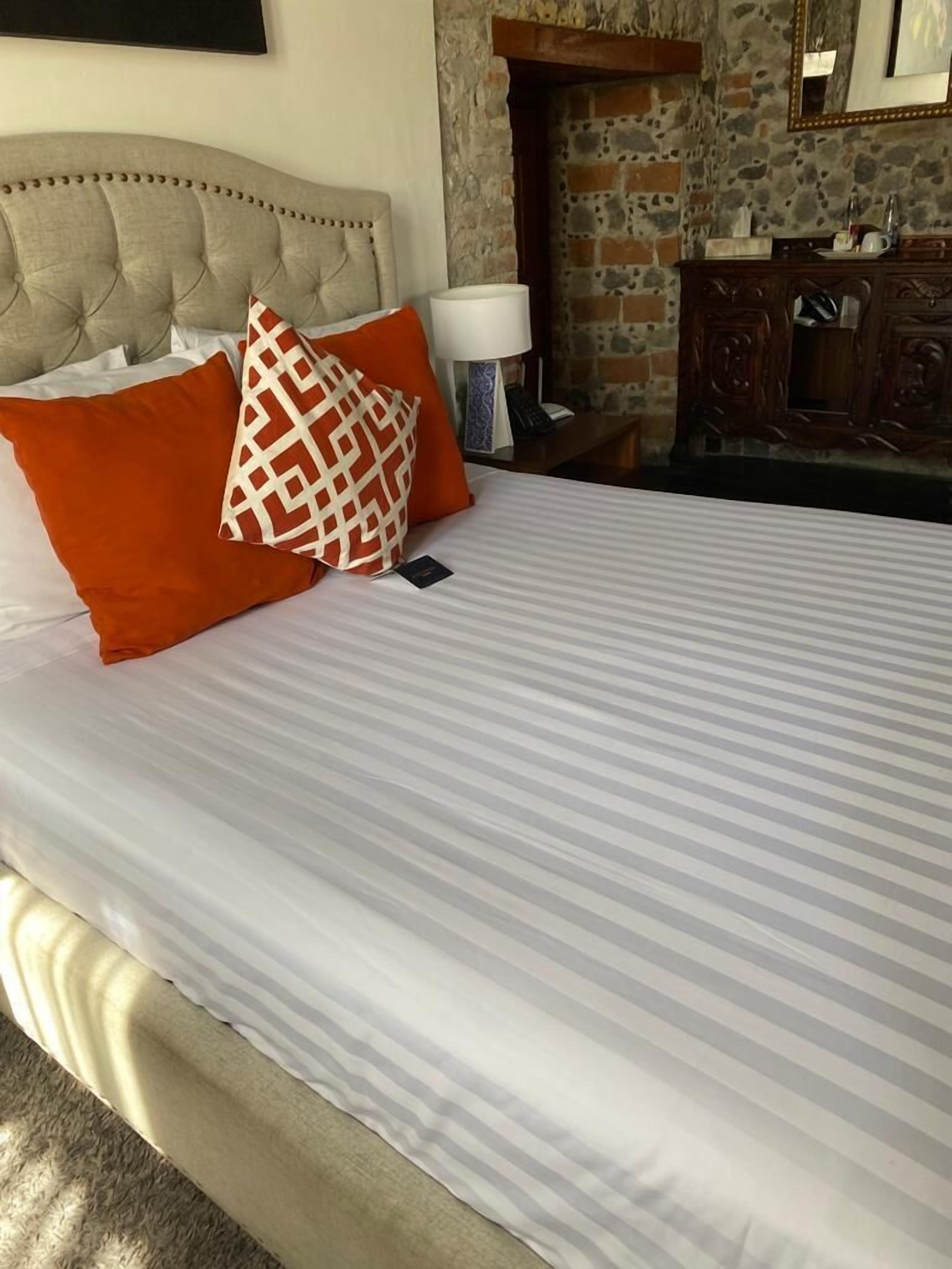 A neatly made bed with striped bedding and two pillows, a bedside lamp, and a wooden dresser in a room with stone walls and a fireplace.