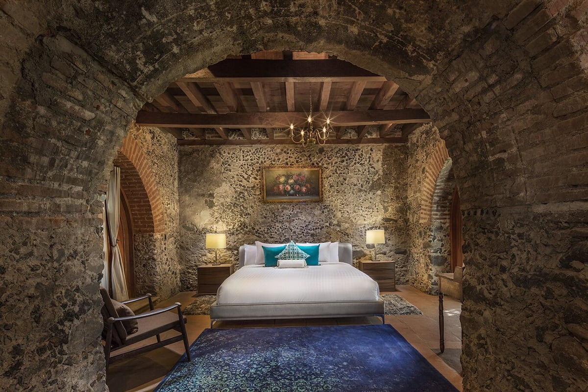 A bedroom with a stone wall, a wooden ceiling, a bed with white bedding, two bedside tables with lamps, and a chandelier.