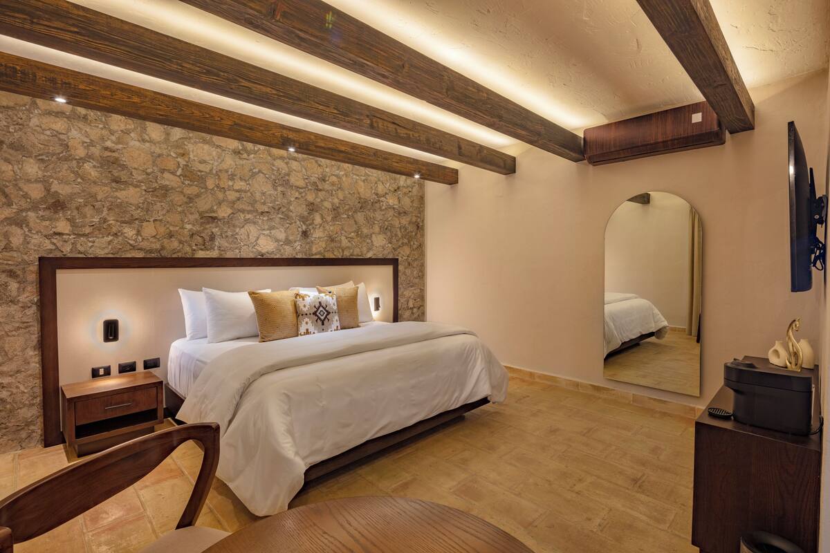 A bedroom with a stone wall, wooden beams, a large bed, bedside tables, a chair, a mirror, and a dresser.