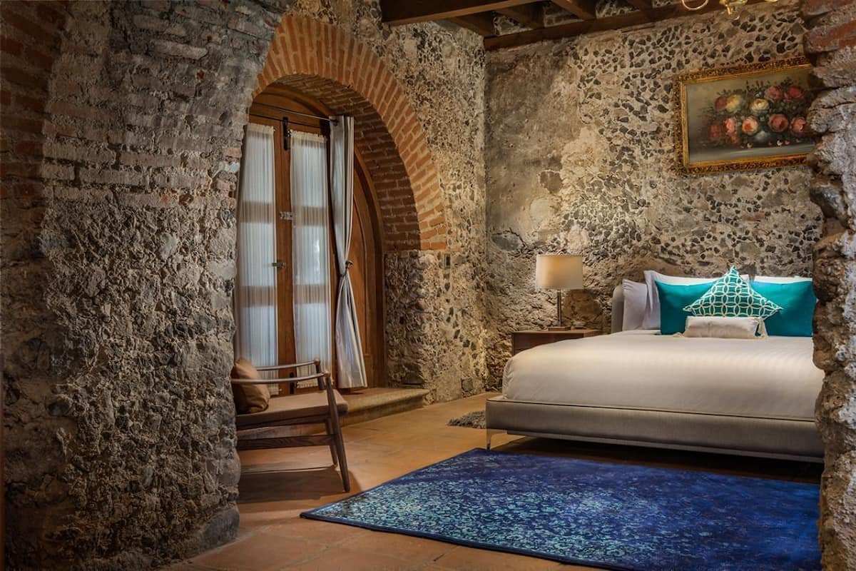 A bedroom with a stone wall, a bed, a nightstand, a chair, and a window with curtains.
