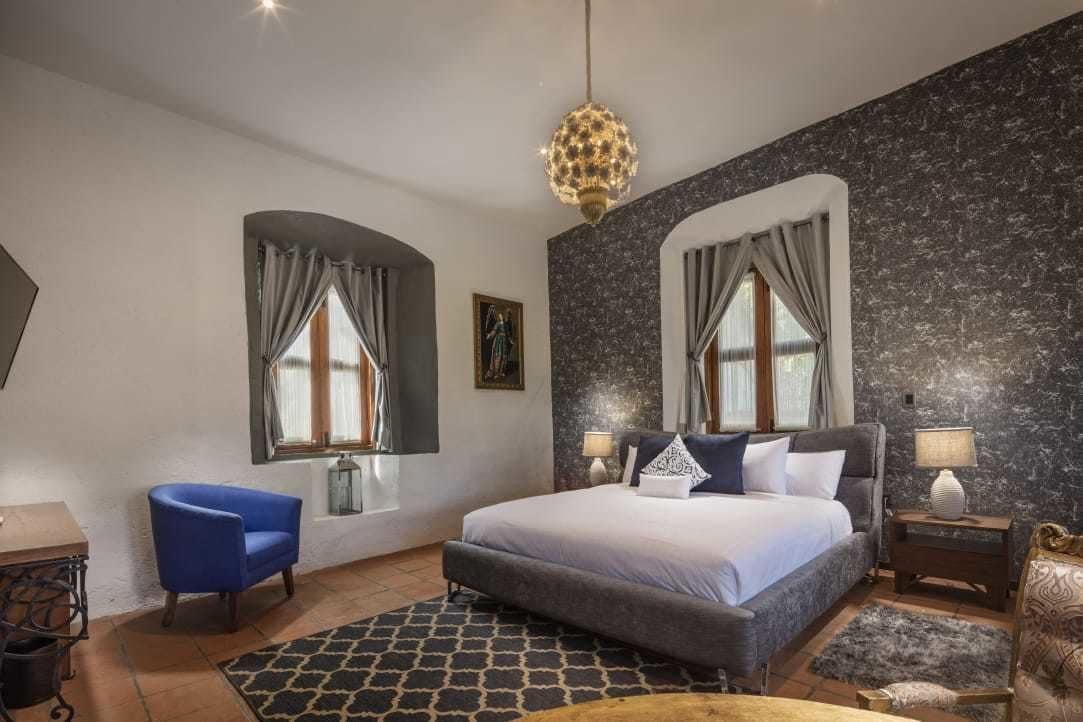 A bedroom with a large bed, two bedside tables, a blue armchair, and a chandelier.