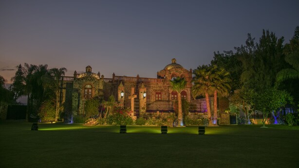 Un edificio grande e histórico con una cúpula y varias torres, rodeado de jardines cuidados y palmeras.
