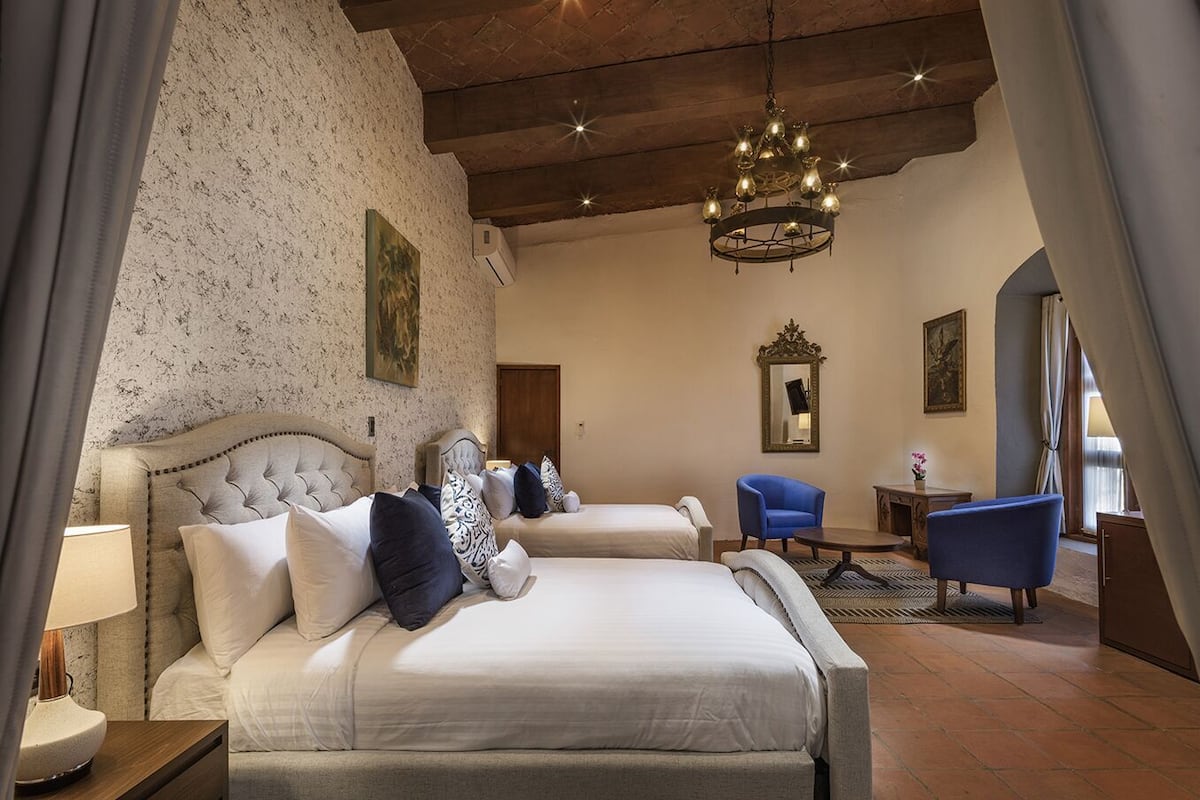 A hotel room with two beds, a chandelier, a blue armchair, and a small table.