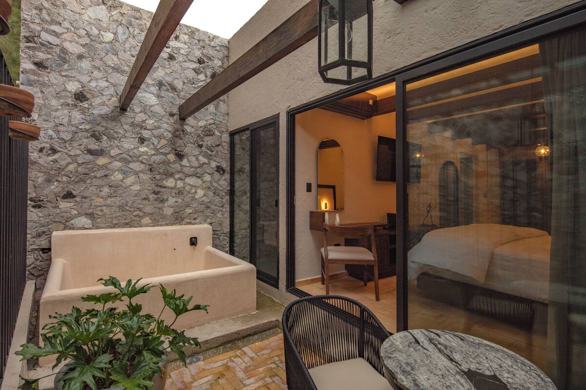 A stone-walled room with a large bathtub, a dining area with a table and chairs, and a bedroom with a bed and a large window.