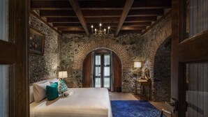 Un dormitorio con pared de piedra, vigas de madera, cama con almohadas, candelabro y ventana con cortinas.
