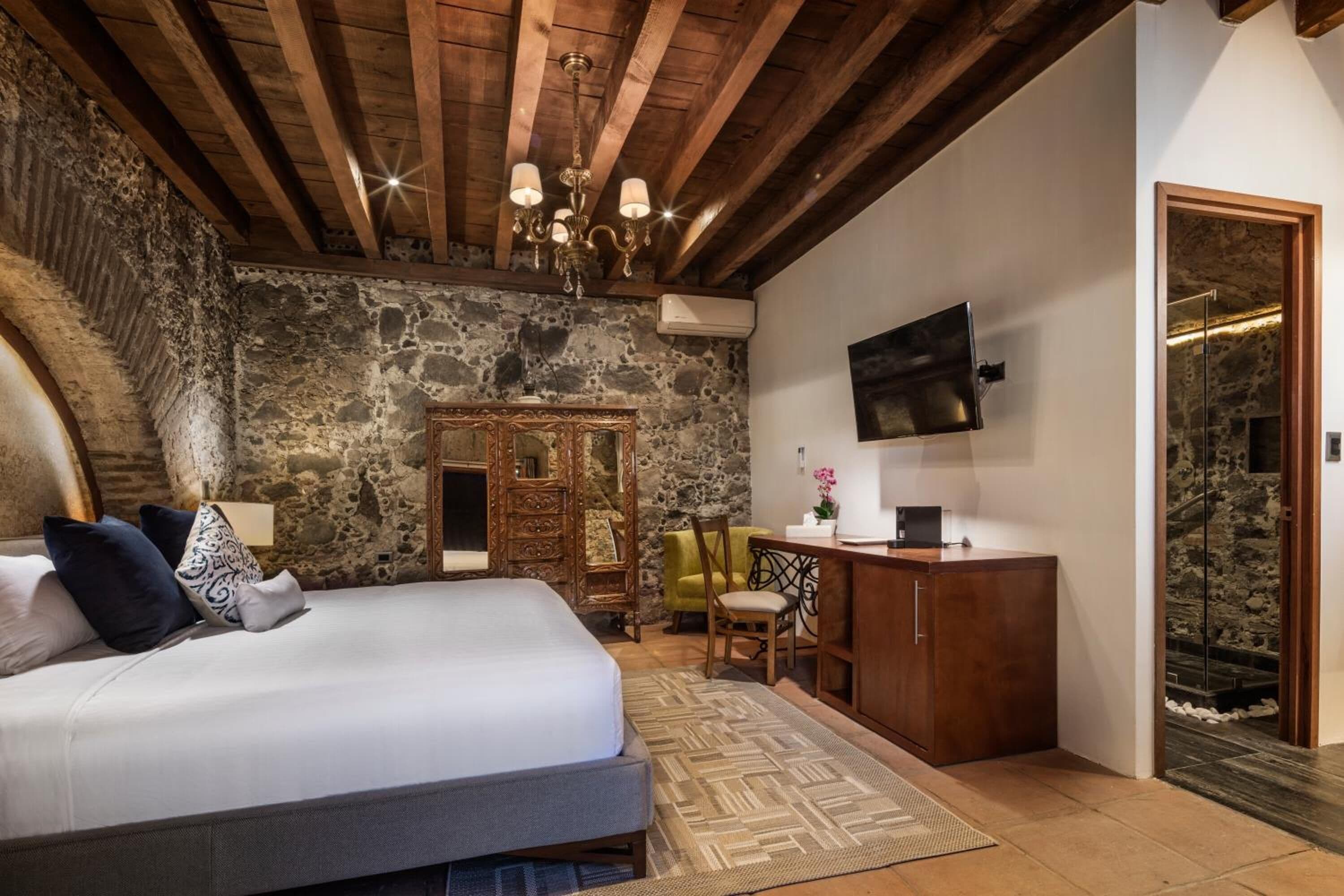 Quarto com parede de pedra, vigas de madeira no teto, cama, escrivaninha, cadeira e televisão na parede.