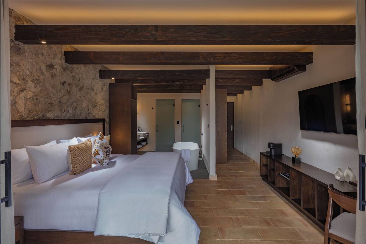 A modern hotel room with a large bed, wooden beams, a stone wall, a bathroom, and a TV mounted on the wall.
