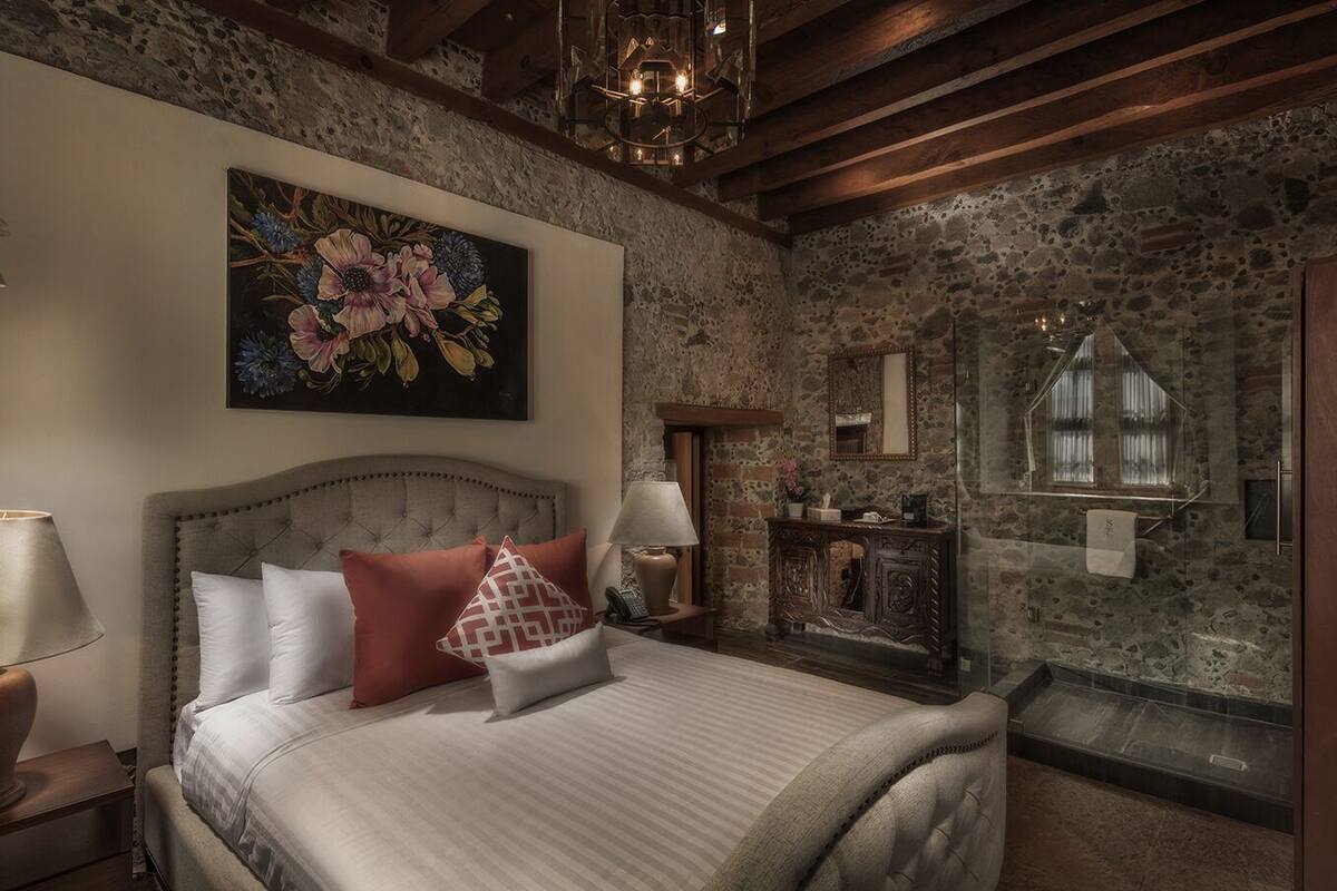 A bedroom with a stone wall, a large floral painting, a bed with white and red bedding, and a wooden headboard.