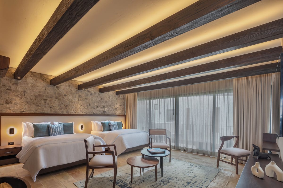 A hotel room with a stone accent wall, a large bed with multiple pillows, two chairs, a small round table, and a flat-screen TV on a wooden stand.