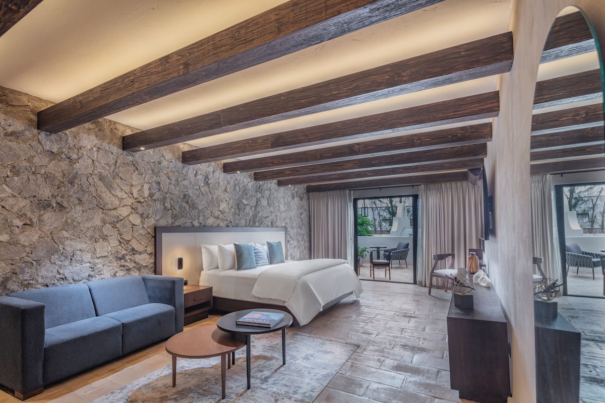 A modern hotel room with a stone wall, a large bed, a sofa, a small table, and a balcony with outdoor seating.