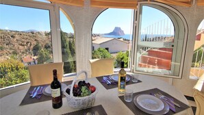 A dining table set with a bottle of wine, a basket of fruit, and a glass of water, overlooking a scenic landscape with buildings and a mountain. - AMIBARCO-Gran Vista-Wifi y Parking Gratis. (Calpe)
