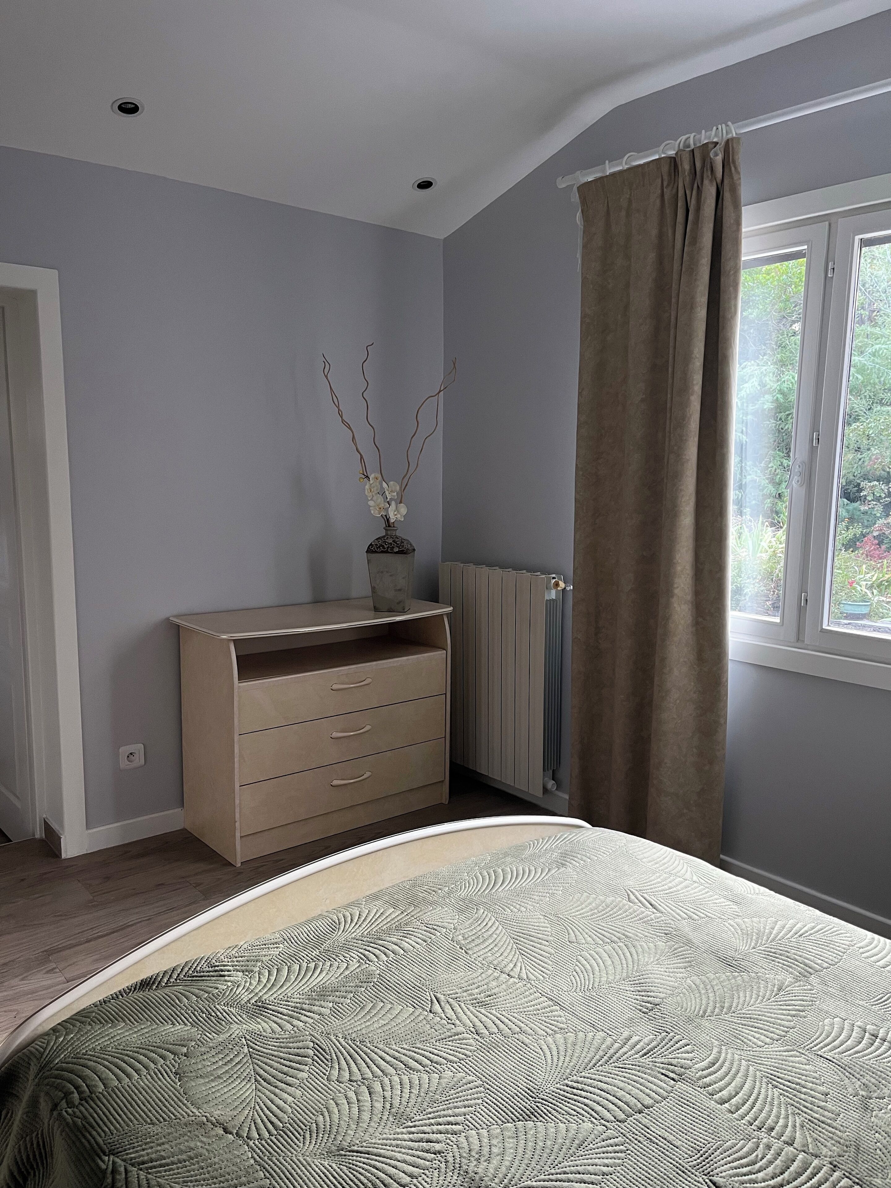 Une chambre à coucher avec un lit, une commode, un radiateur et une fenêtre avec des rideaux.