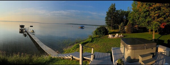 A lakeside deck with a hot tub, a dock with a boat, and a grassy area with chairs.