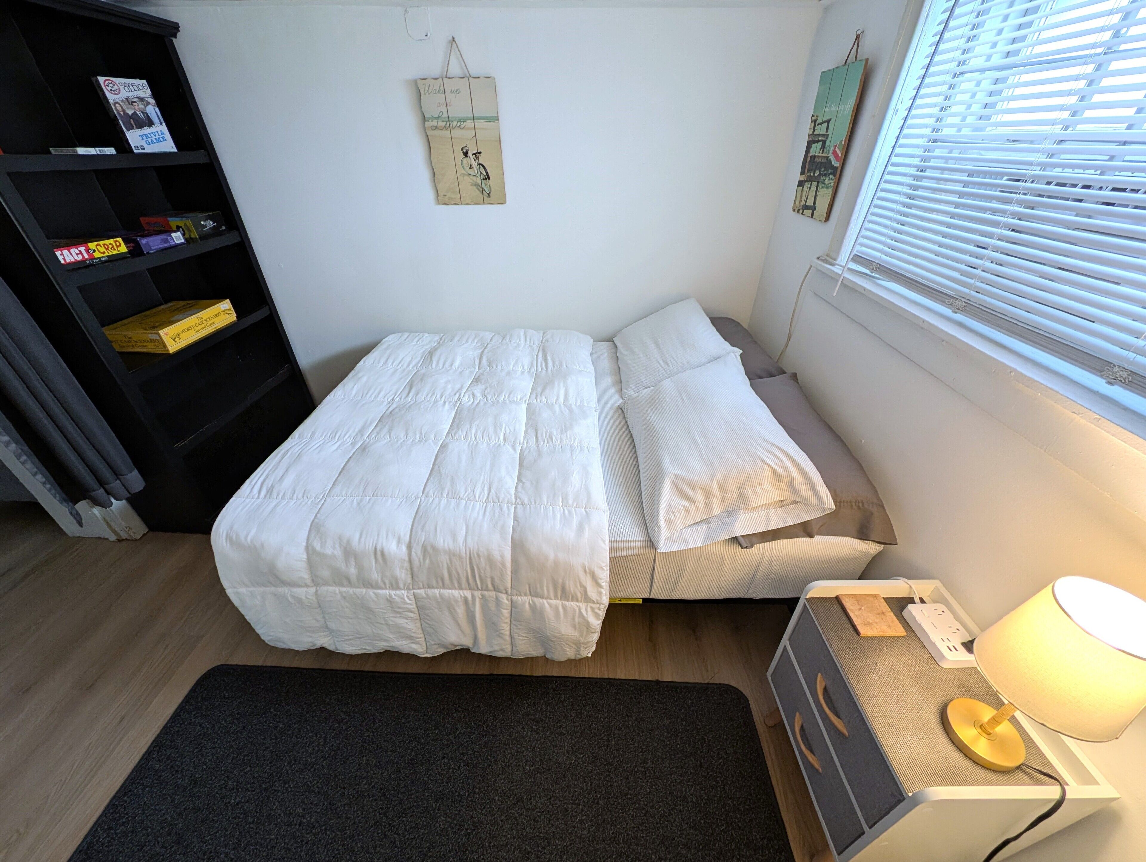 A neatly made bed with a white duvet and pillows, a small bedside table with a lamp, a bookshelf with various items, and a window with blinds.