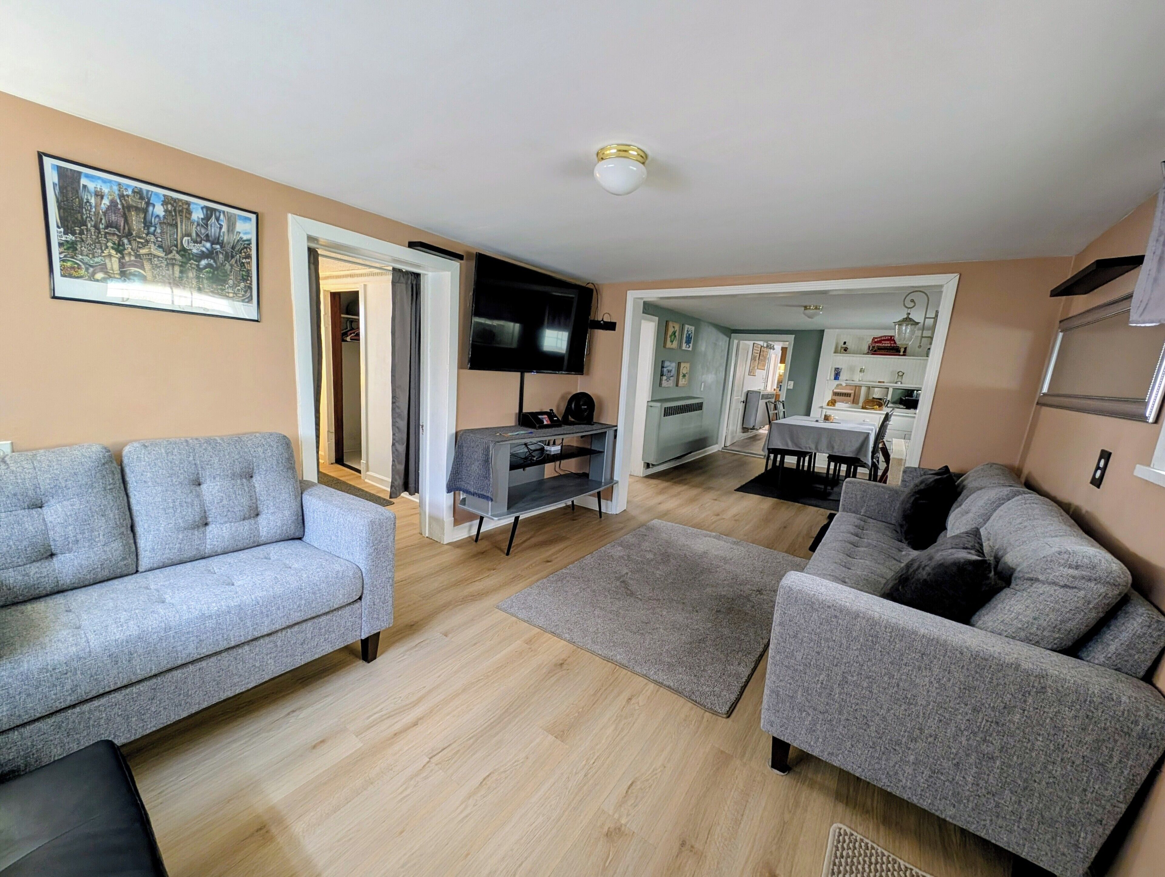 Een moderne woonkamer met grijze banken, een flatscreen televisie en een eethoek met een tafel en stoelen.