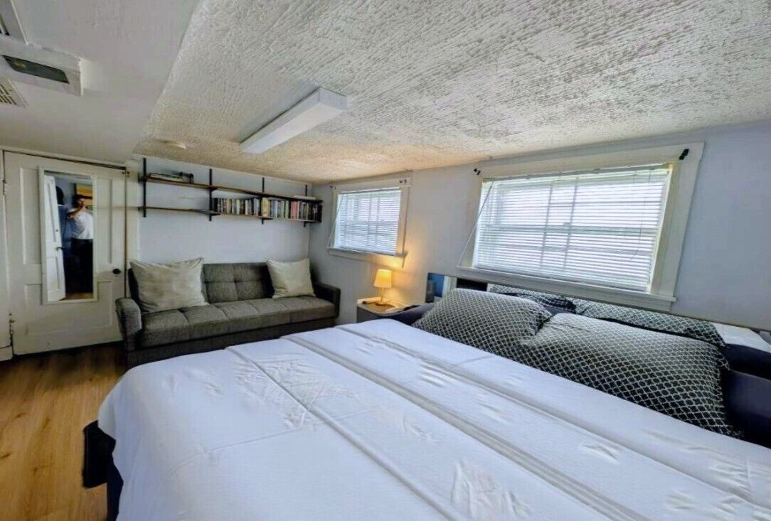 A bedroom with a large bed, a sofa, a bedside table with a lamp, a bookshelf, and a door leading to another room.