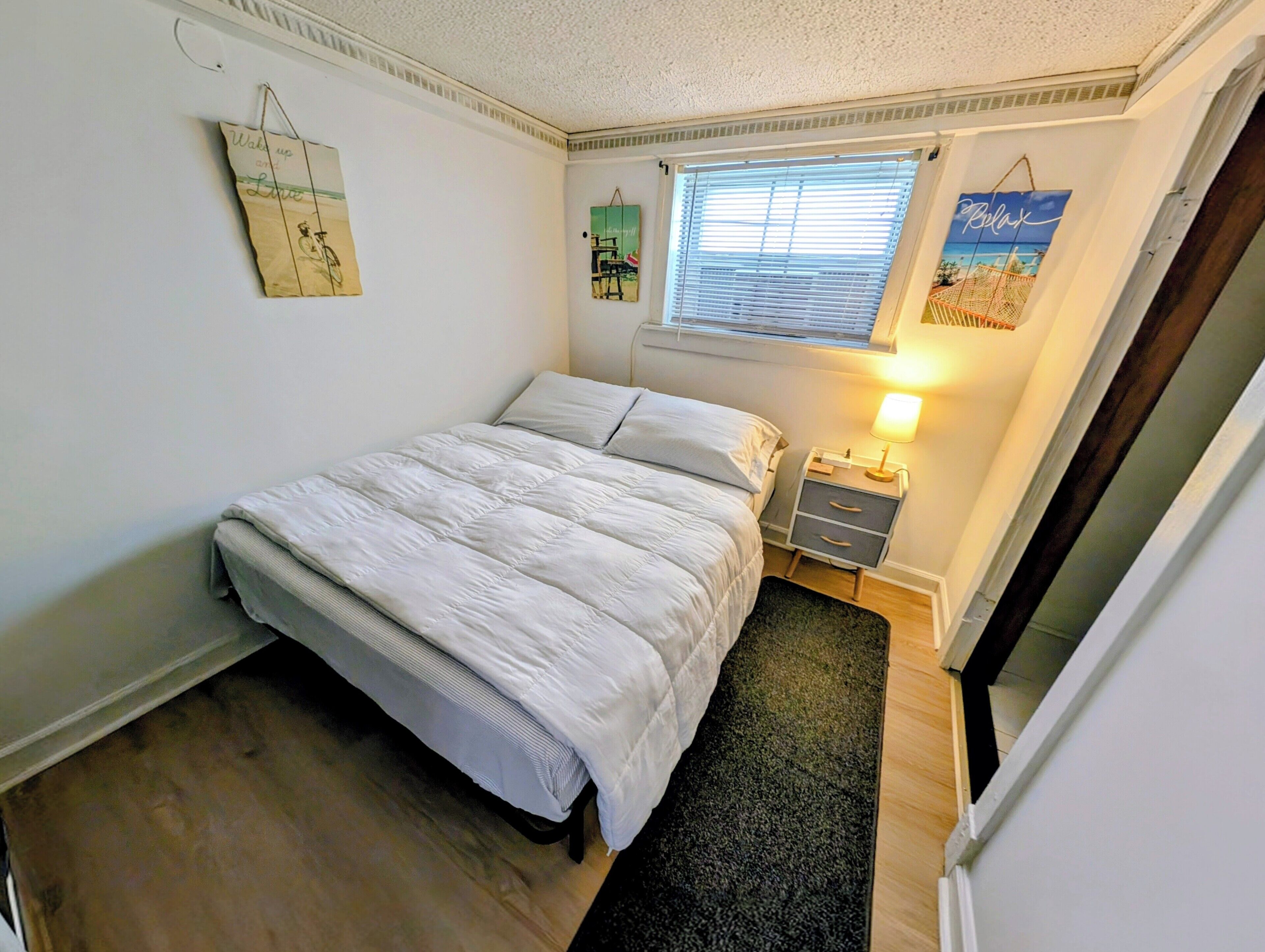A bedroom with a bed, a nightstand with a lamp, a window with blinds, and wall decorations.