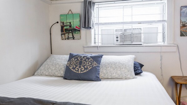 Un dormitorio con cama, almohadas, una ventana con persianas, un radiador y una obra de arte colgada en la pared.