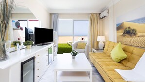 Een moderne woonkamer met een flatscreen-tv, een witte salontafel, een gele bank en een groot raam met uitzicht op zee.
