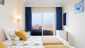 A hotel room with a bed, a desk, a chair, a balcony with a view, and a painting on the wall.