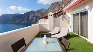 A balcony with a table, chairs, and a lantern. It overlooks a body of water and a mountainous landscape.