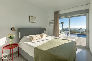 A modern bedroom with a large bed, a red side table, a vase of flowers, and a view of a pool and ocean. - Villa NEVIS by Abahana Villas (Benitachell)