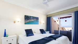 Une chambre d’hôtel avec deux lits, un tableau représentant une plage, un balcon avec des rideaux bleus et une table de chevet blanche ornée d’un vase.