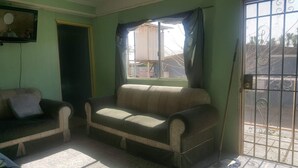 Un salon avec un canapé et un fauteuil, un téléviseur et une fenêtre avec des rideaux.