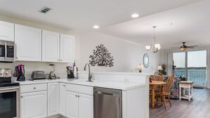 A modern kitchen with white cabinets, stainless steel appliances, and a dishwasher. There is a dining area with a table and chairs, and a view of the ocean through large windows. - Luxury condo with amazing bay front view (Ocean City)