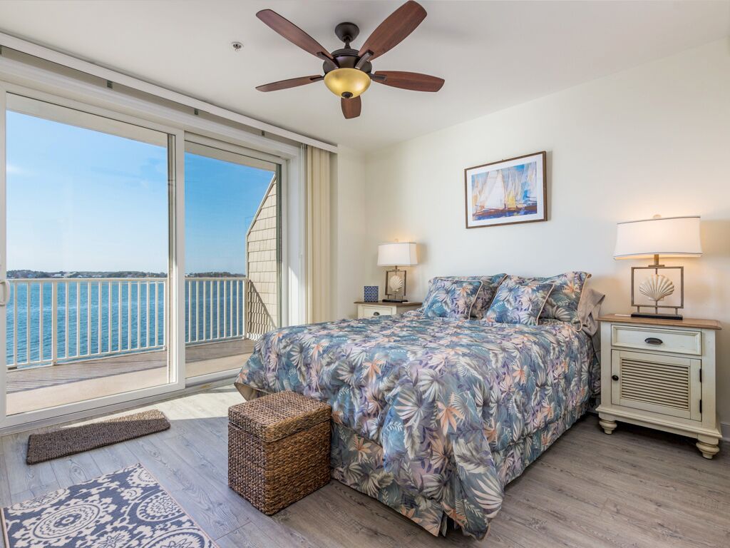 A bedroom with a large window, a ceiling fan, a bed with floral bedding, a nightstand, and a view of the sea.