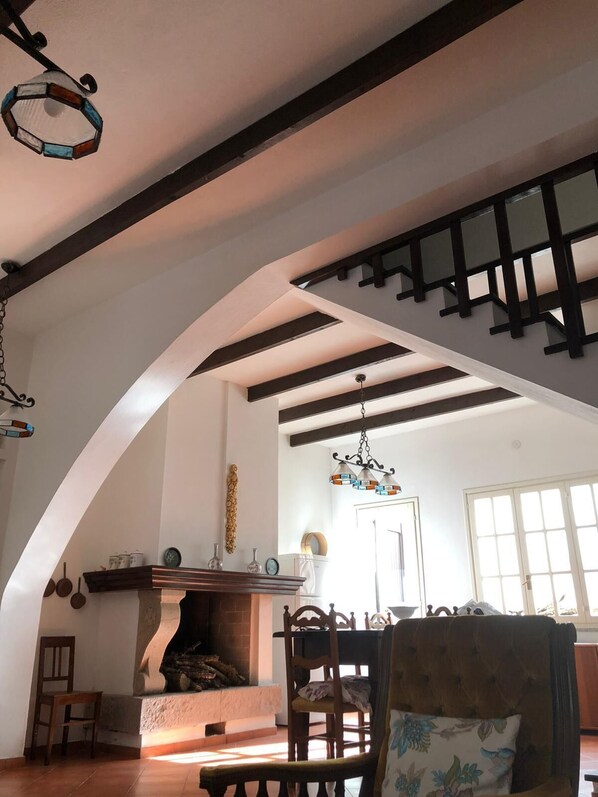A spacious living room with a high ceiling, exposed wooden beams, a fireplace, and a dining area with a wooden table and chairs.
