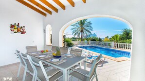 Un coin repas avec vue sur la piscine, comprenant une table mise avec des assiettes, des verres et un panier de fruits.