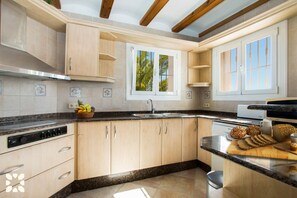 A kitchen with wooden cabinets, a granite countertop, a built-in oven, and a window with a view of palm trees. - Villa SOLEADA by Abahana Villas (Calpe)
