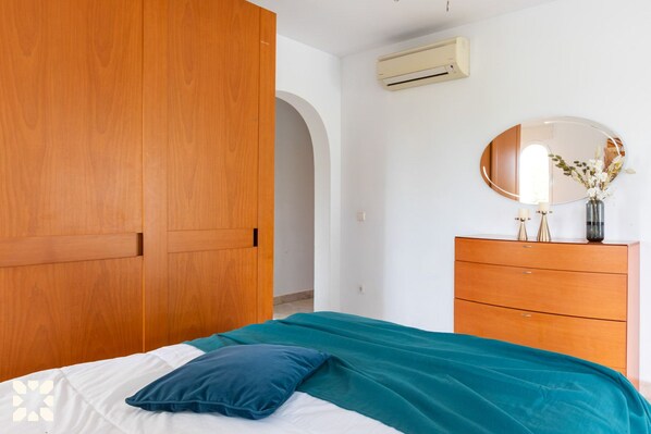 A bedroom with a wooden wardrobe, a bed with teal bedding, a dresser with a mirror, and an air conditioning unit on the wall.